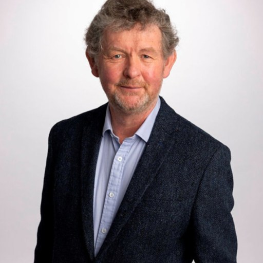 An older man with curly gray hair, smiling slightly, wearing a blue blazer and gray shirt, standing against a light background.