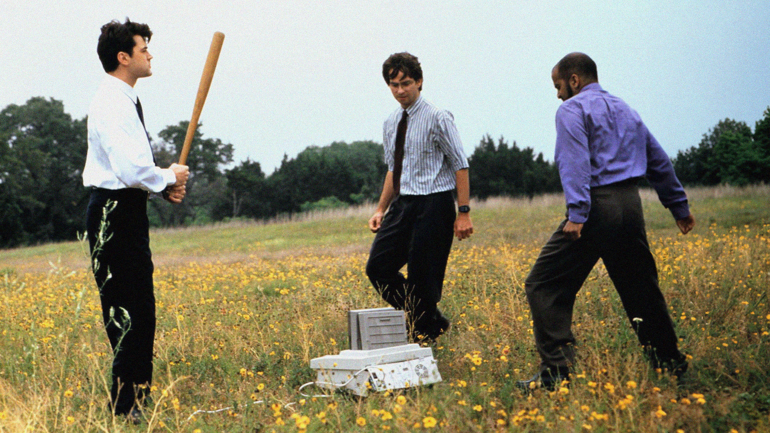Three men in business attire standing in a field, one holding a baseball bat, next to a damaged printer or computer.