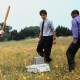 Three men in business attire standing in a field, one holding a baseball bat, next to a damaged printer or computer.