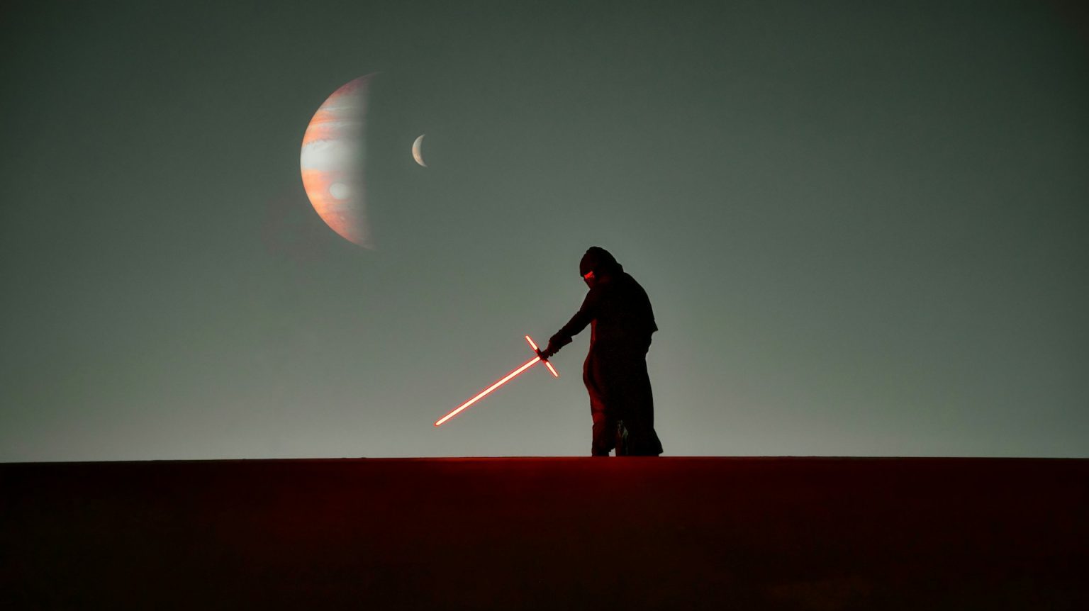 A person in a dark robe wielding a glowing red lightsaber on a desert dune under a night sky with a large crescent moon and a visible planet.