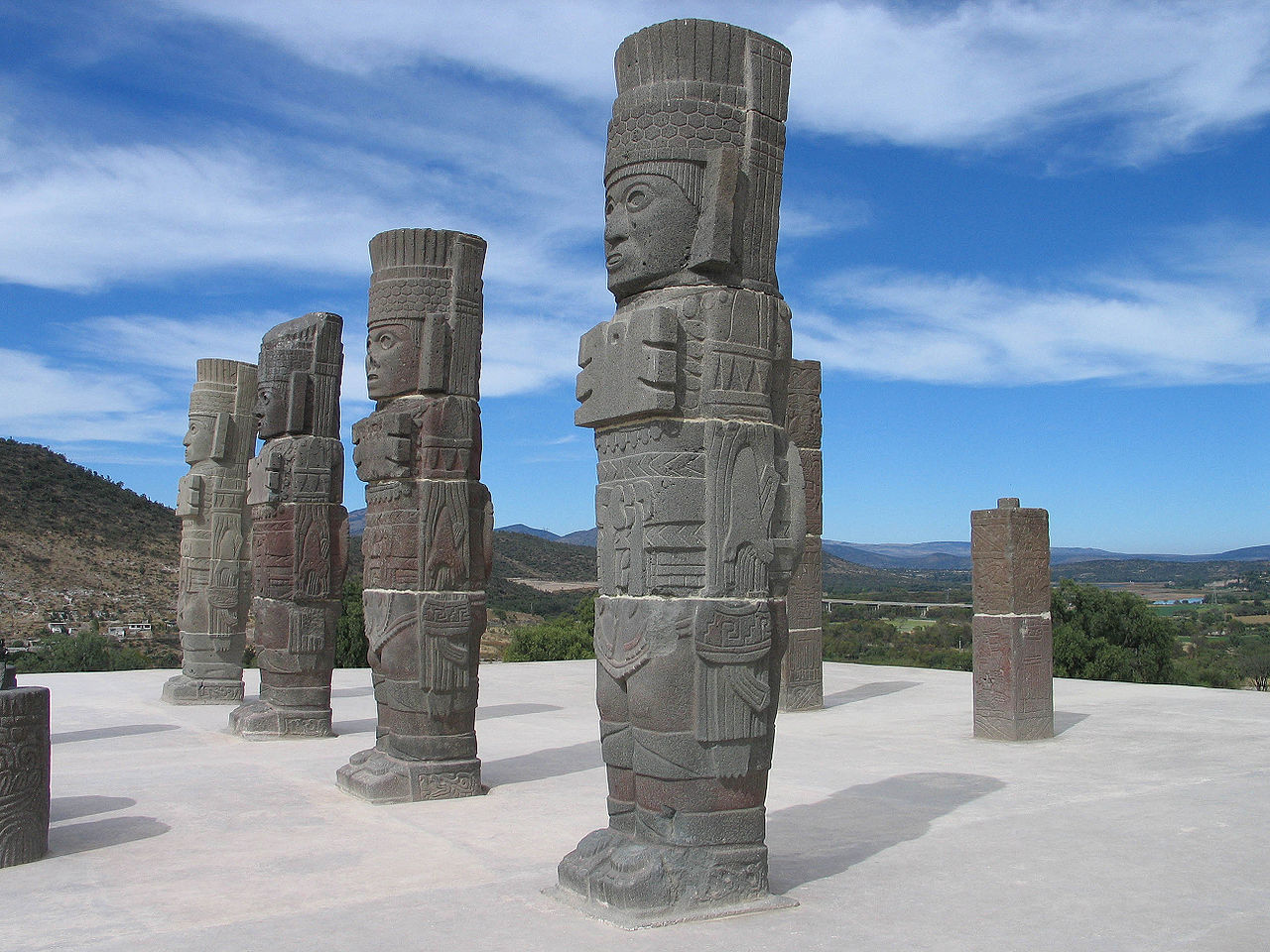 Stone statues of ancient Toltec warriors stand in a row on a platform with a scenic landscape in the background at Tula Archaeological Site, Mexico.