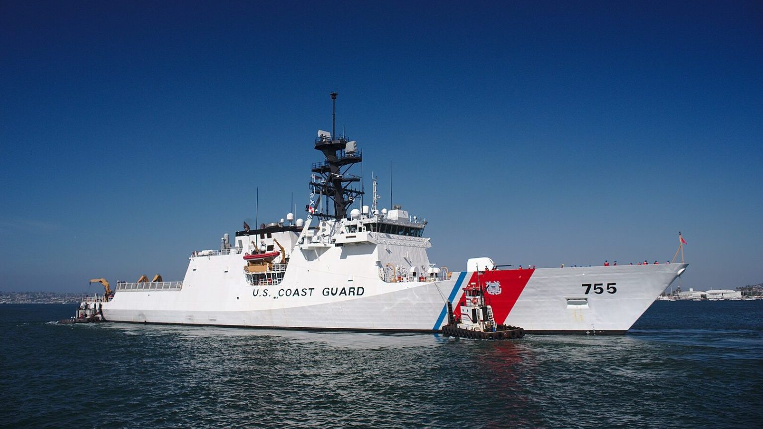 A u.s. coast guard cutter sails on calm blue waters against a clear sky, flying the coast guard flag on its stern.