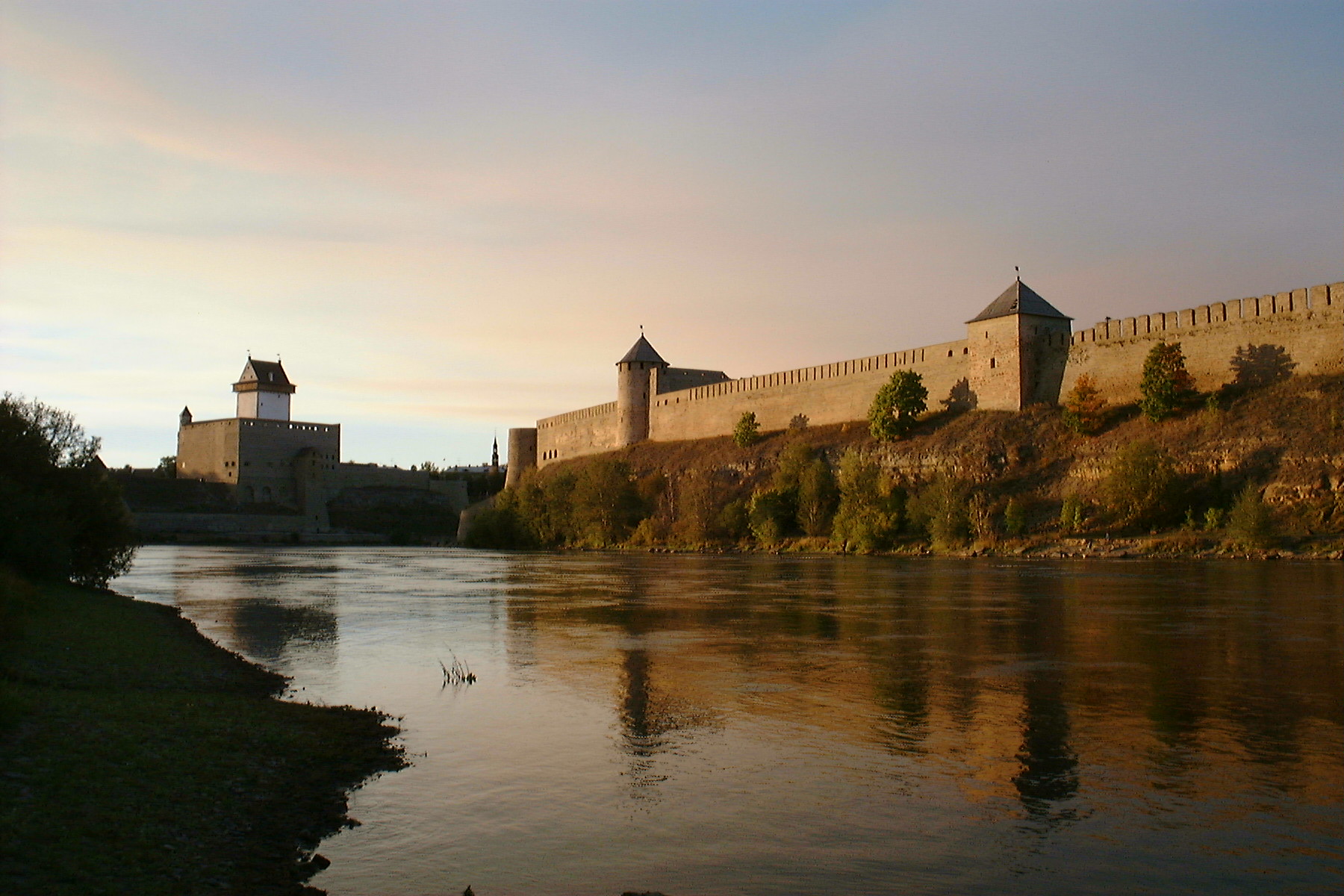 A river runs alongside an old stone fortress with multiple towers and walls, set against a sunset sky. Trees and greenery line the opposite bank.