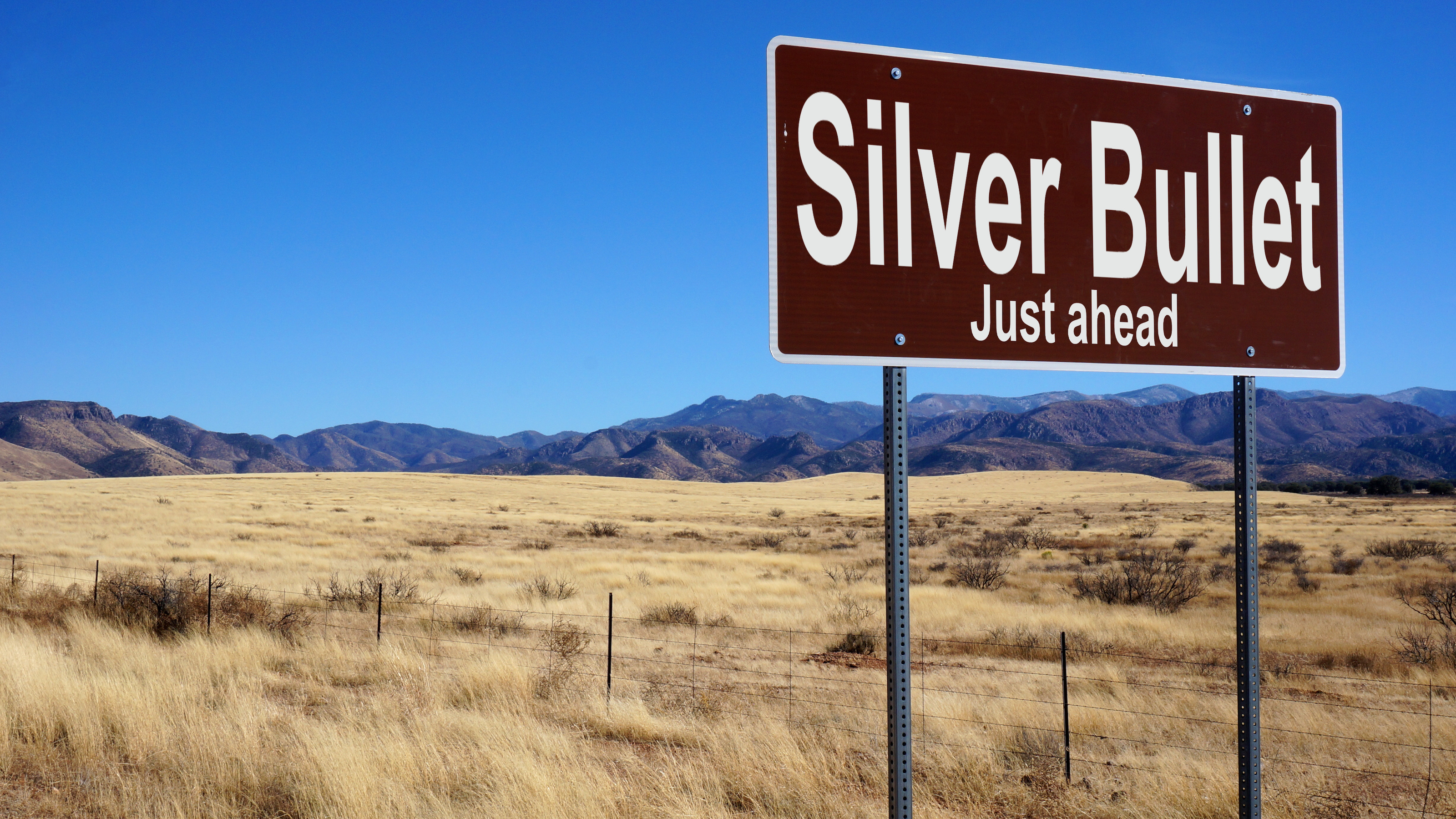 A brown road sign reads "Silver Bullet Just ahead" against a backdrop of dry grassy plains and distant mountains under a clear blue sky.