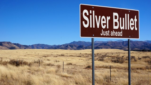 A brown road sign reads "Silver Bullet Just ahead" against a backdrop of dry grassy plains and distant mountains under a clear blue sky.
