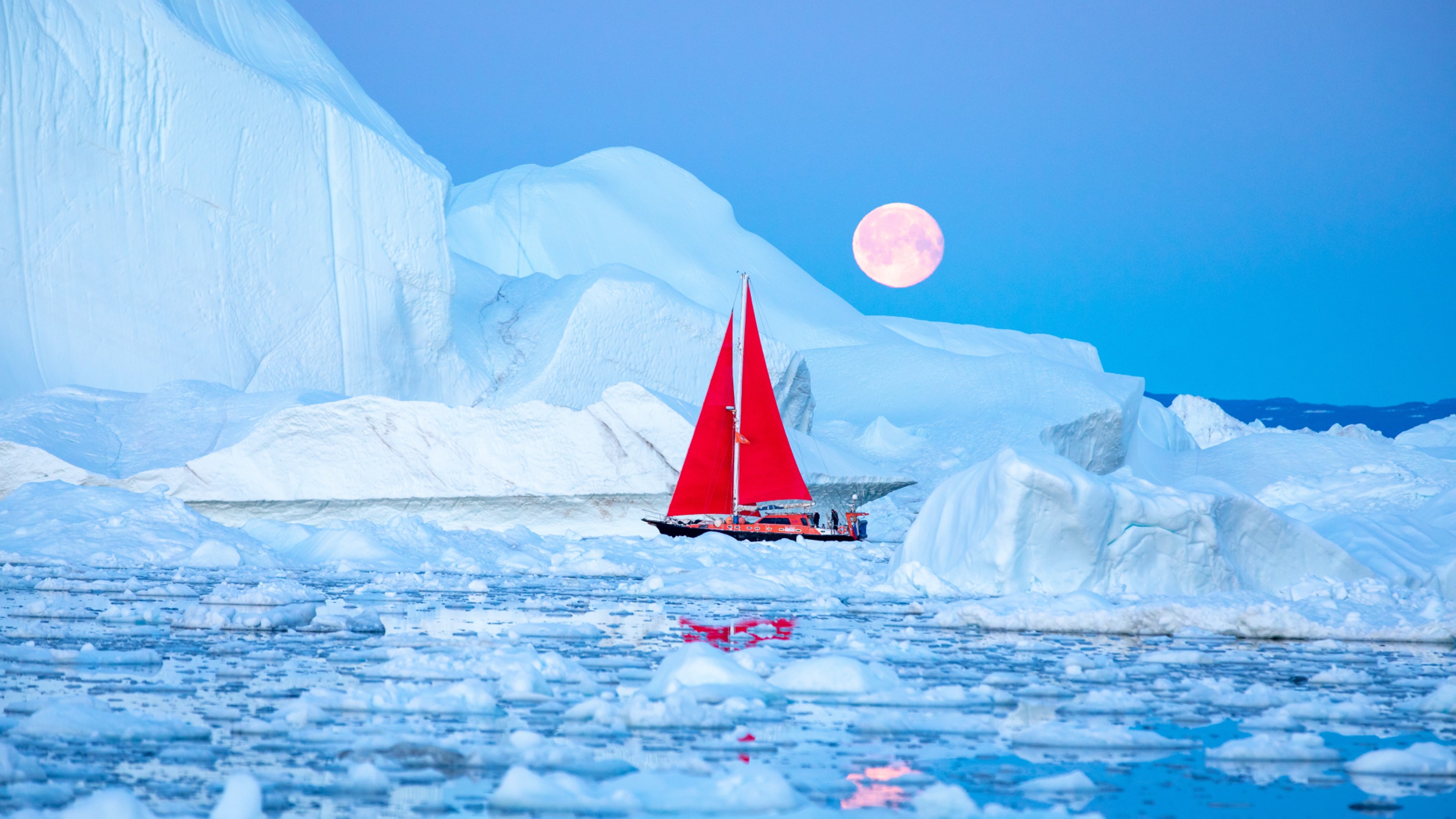 A sailboat with a red sail navigates through icy waters surrounded by large icebergs under a full moon.
