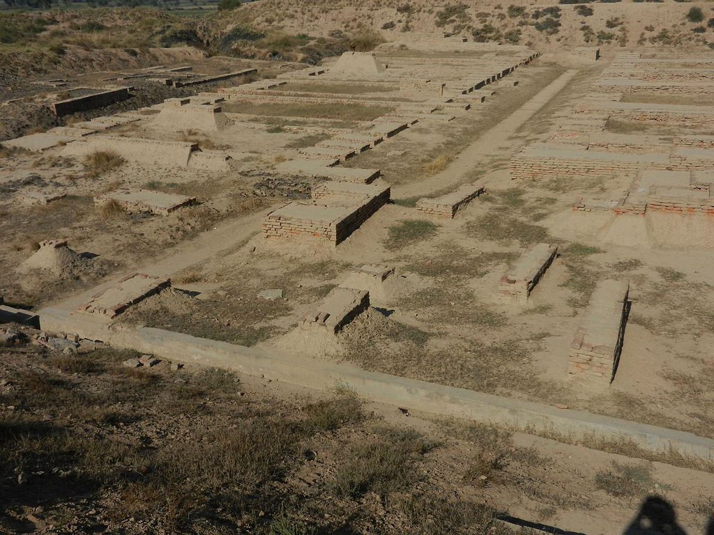 Archaeological site featuring ancient ruins, with rectangular brick structures partially buried in dirt and sand, surrounded by a dry, barren landscape.
