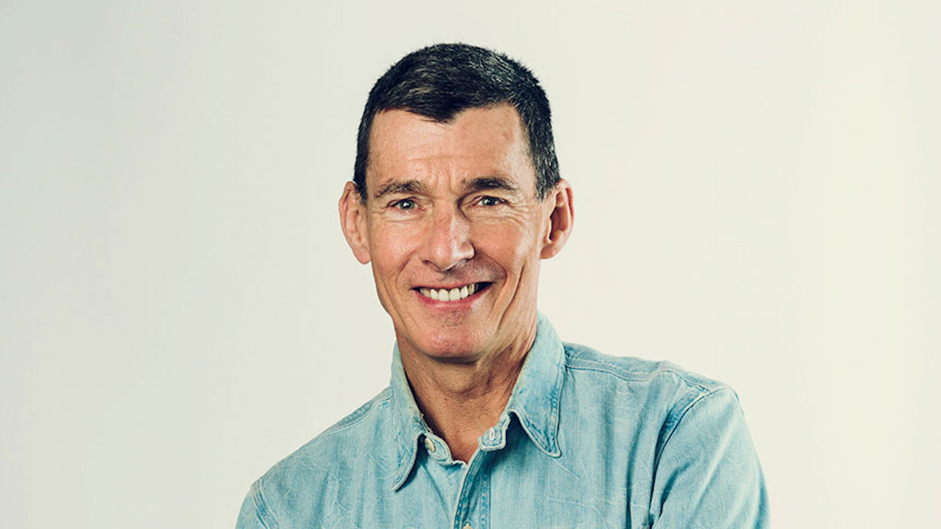 A middle-aged man with short dark hair, wearing a light blue denim AI shirt, smiling at the camera against a plain light background.