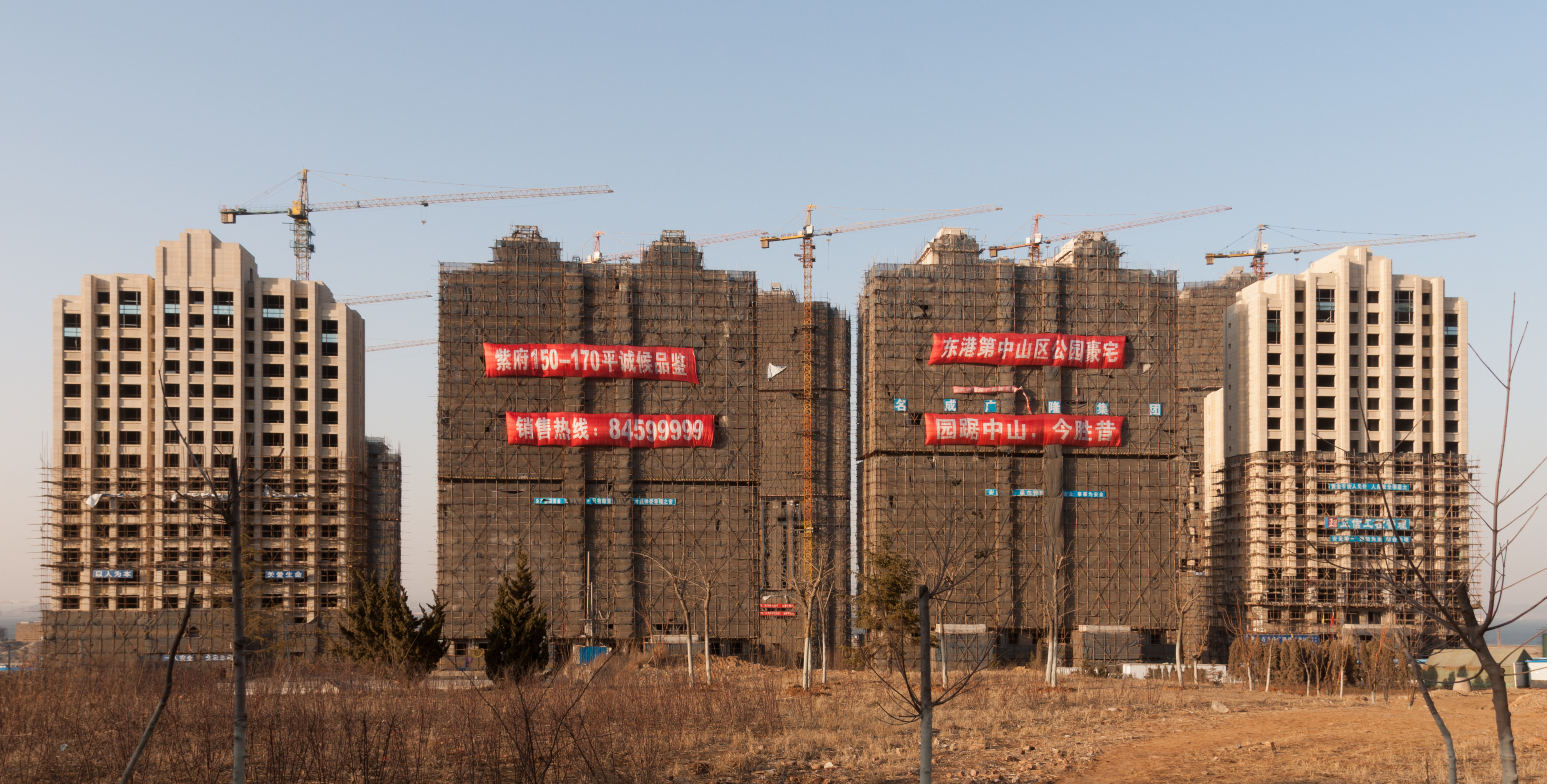 Four high-rise buildings under construction with scaffolding and cranes, featuring large red banners with text in Chinese, situated in a barren area with scattered trees and a clear sky.