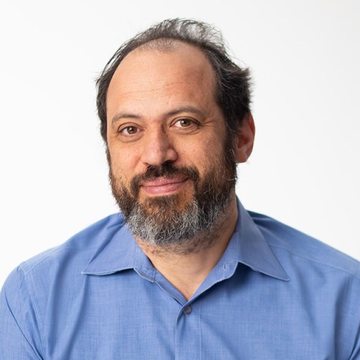 A middle-aged man with a beard and thinning hair, wearing a blue button-up shirt, looks at the camera against a plain white background.