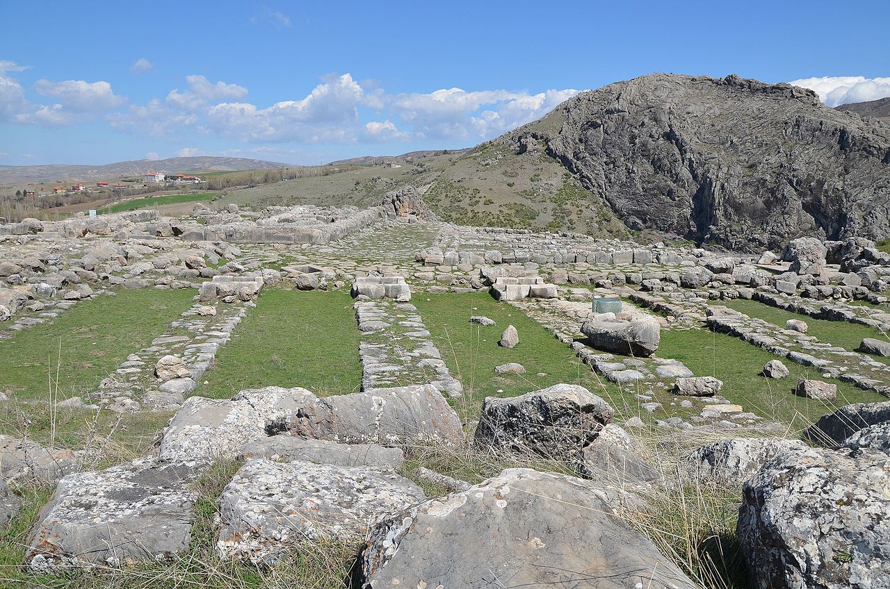 Ancient ruins with stone foundations, columns, and scattered rocks set against a backdrop of hills and a partly cloudy sky.