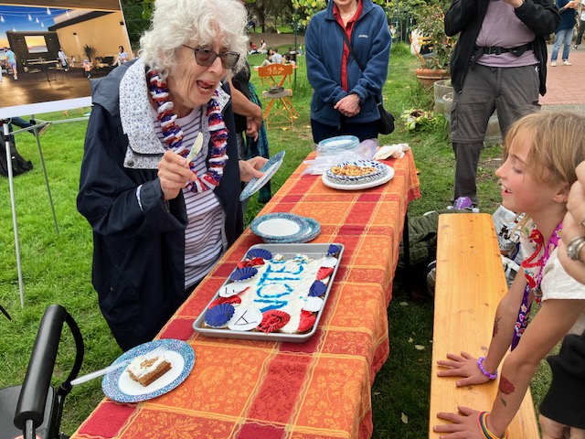 An elderly woman with glasses smiles at a child across a table with patriotic-themed cakes during an outdoor event.