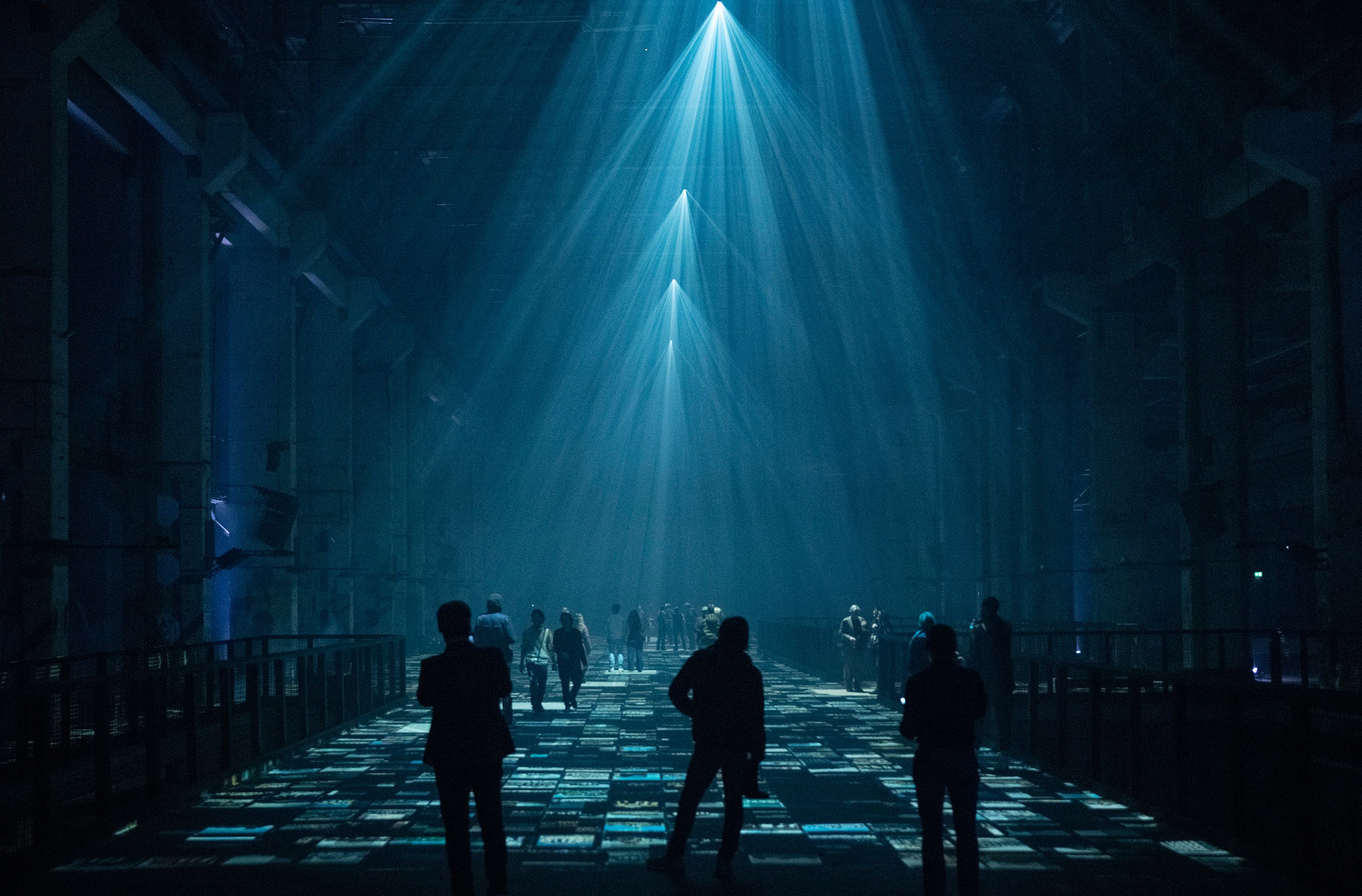 A group of people standing in a large, dimly-lit industrial space with beams of light projecting downwards, creating geometric patterns on the floor.