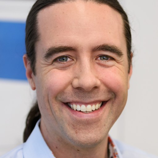 A smiling person with long dark hair tied back, wearing a light blue shirt, in a close-up shot.