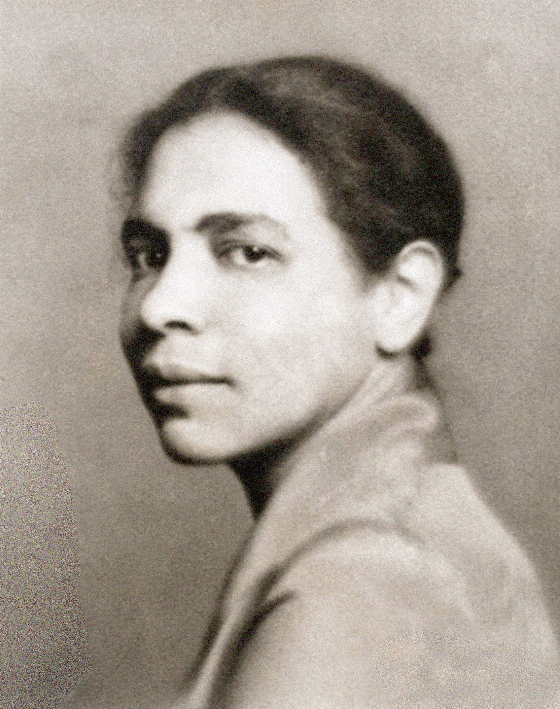 Black and white portrait of a person with short, dark hair, wearing a light-colored garment, looking over their left shoulder directly at the camera. The background is plain and blurred.