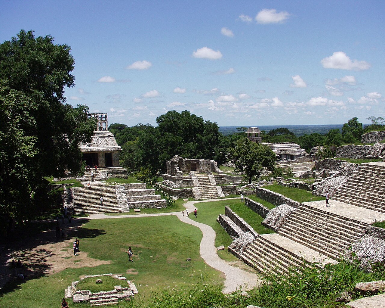 View of ancient Mayan ruins with stone steps, platforms, and structures surrounded by lush greenery and pathways. Visitors are walking around the archaeological site under a partly cloudy sky.