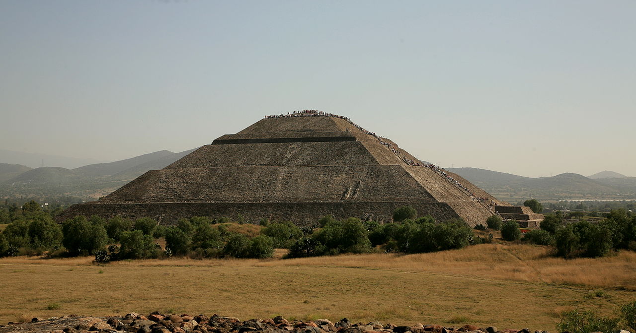A large, ancient stone pyramid sits against a clear sky, surrounded by sparse vegetation and a dry, grassy landscape. People are seen ascending its steep steps.