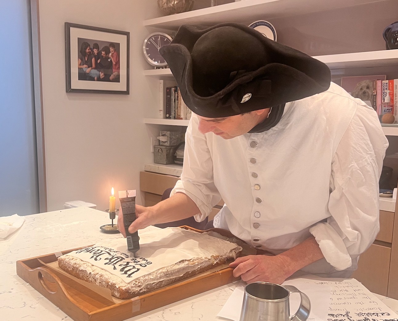 A man in traditional clothing lighting a candle on a rectangular cake at a kitchen table, with family photos and a clock in the background.