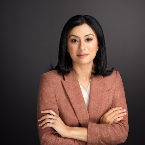 A woman in a rust-colored blazer stands with her arms crossed against a dark gray background, looking directly at the camera.