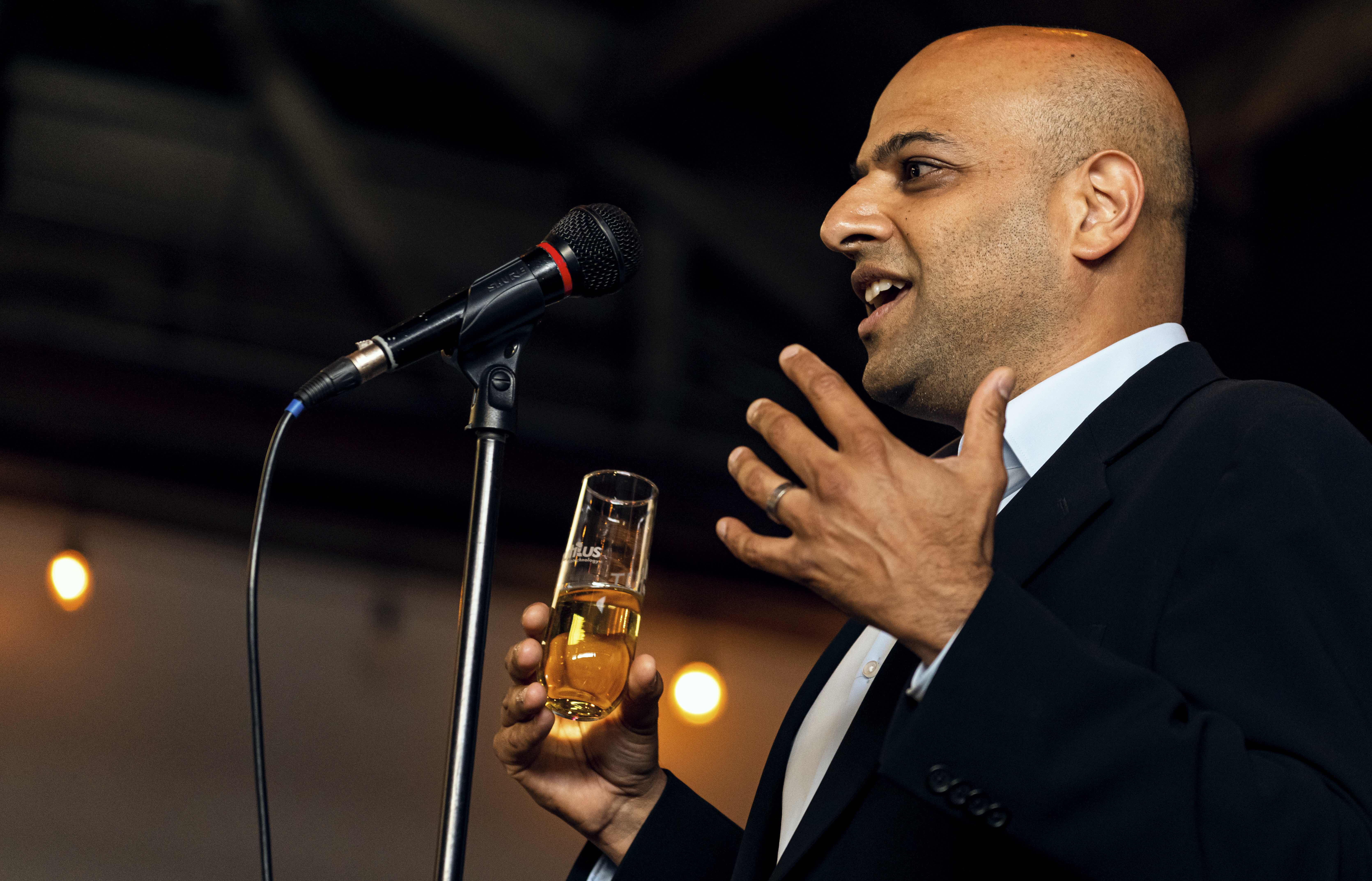 A man in a suit speaks into a microphone while holding a glass of champagne.