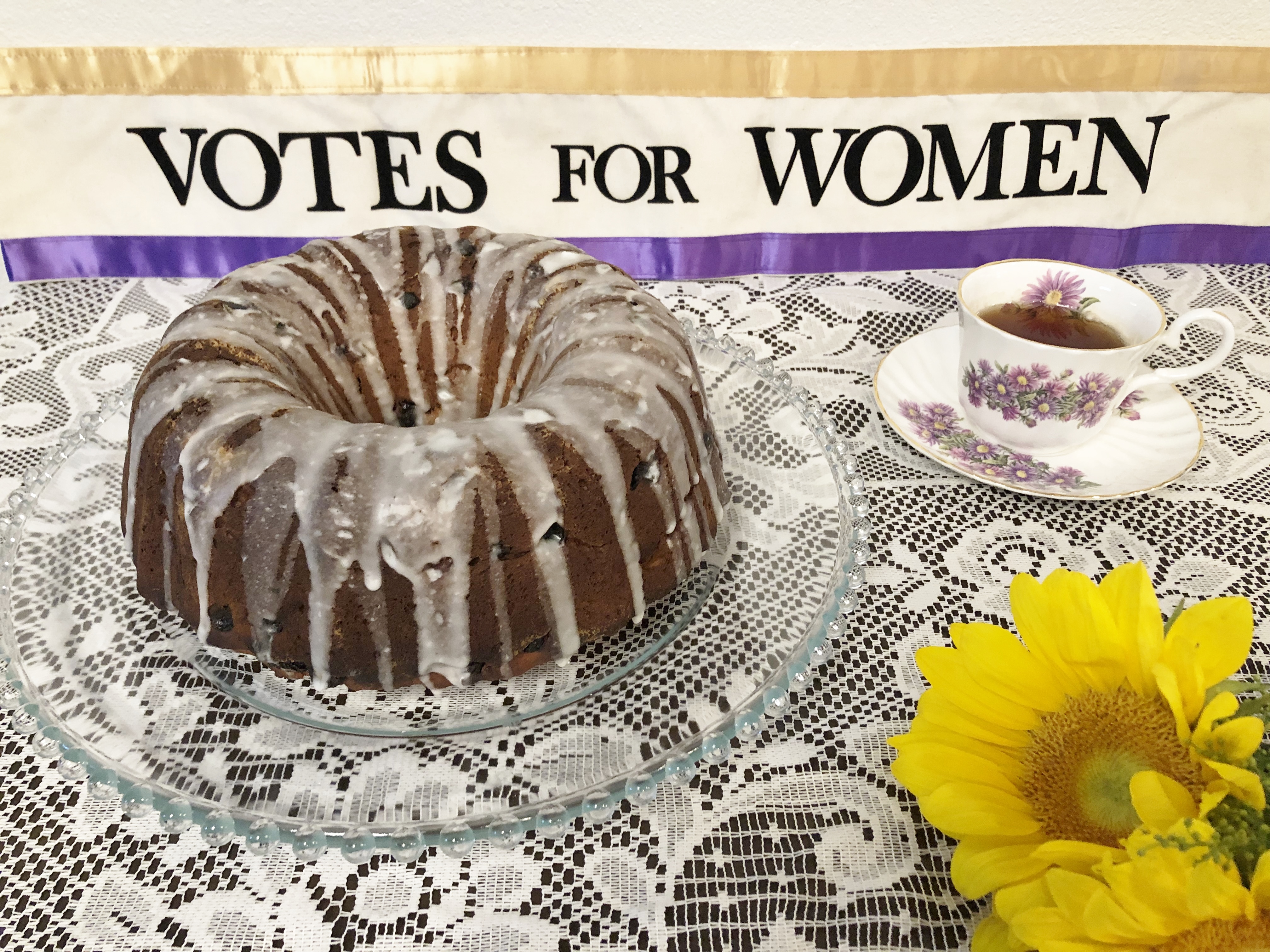 A bundt cake with white glaze on a glass stand, a cup of tea on a saucer, and a sunflower, under a "votes for women" banner.