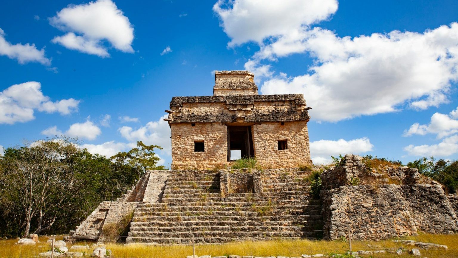 Ancient stone structure with stairs under a blue sky with scattered clouds, surrounded by vegetation.