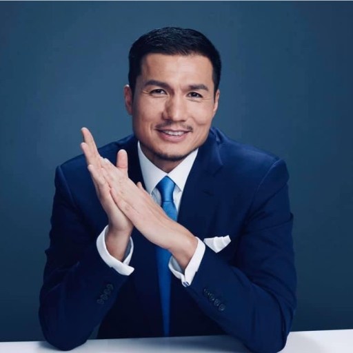 A man in a blue suit and tie sits at a table, smiling with his hands clasped in front of him.