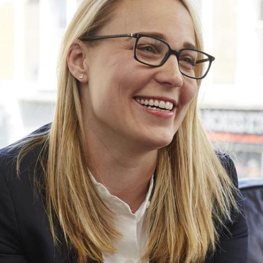 A smiling woman with blonde hair and glasses, wearing a navy blazer and white shirt, seated indoors.