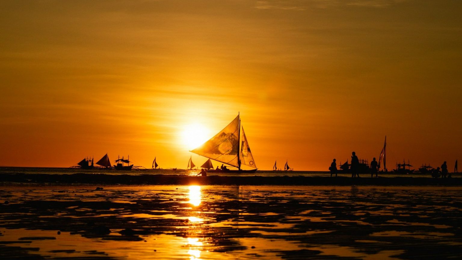 A sailboat is silhouetted against a vibrant sunset over a calm sea, with other boats and their reflections visible in the background. The sky and water have a golden hue.