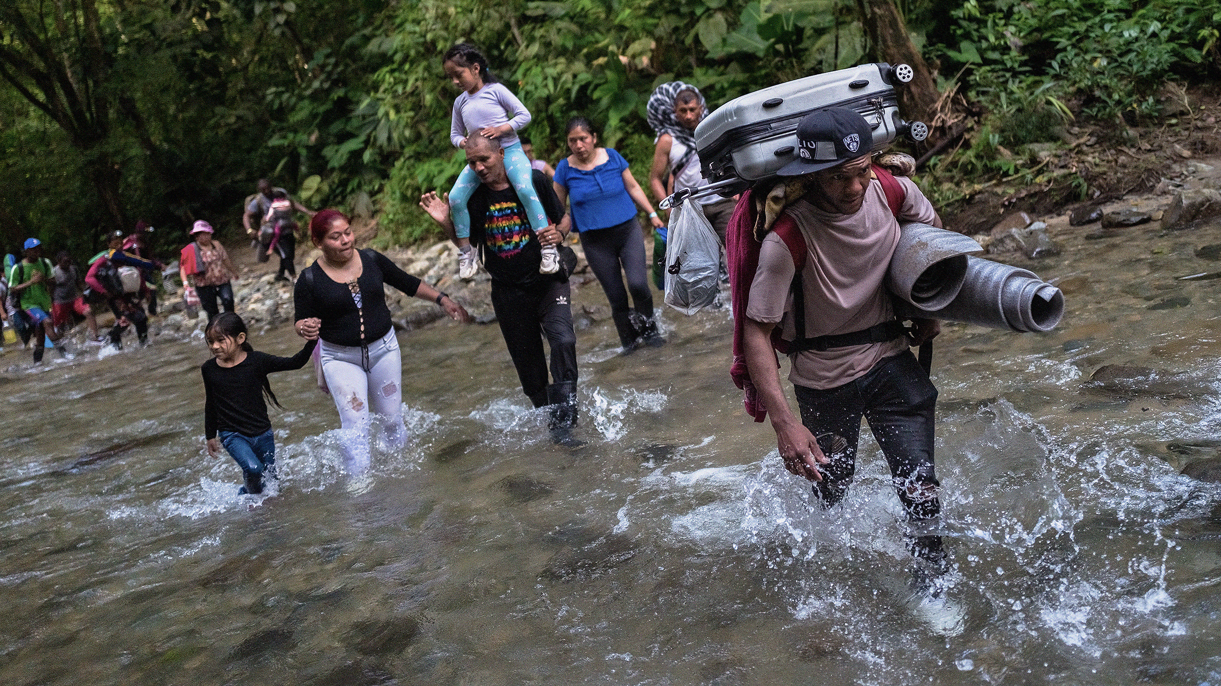 A group of people, including children, wade through a shallow river in the forested area of the Darién Gap. One person carries a suitcase and other luggage on their back.