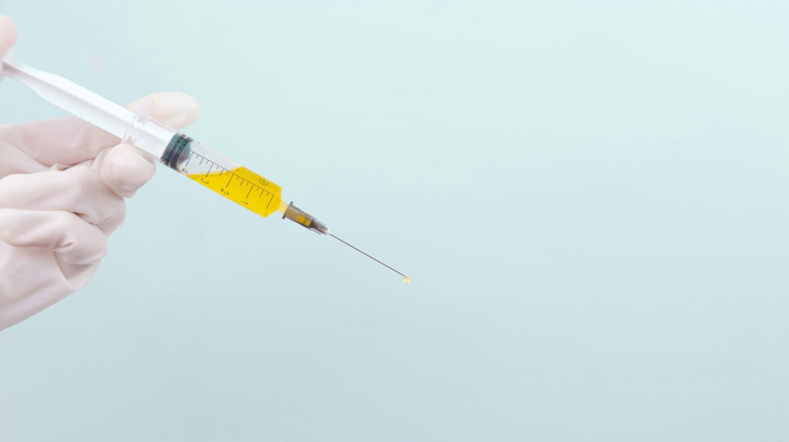 A hand in surgical gloves holding a syringe filled with yellow fluid against a light blue background.