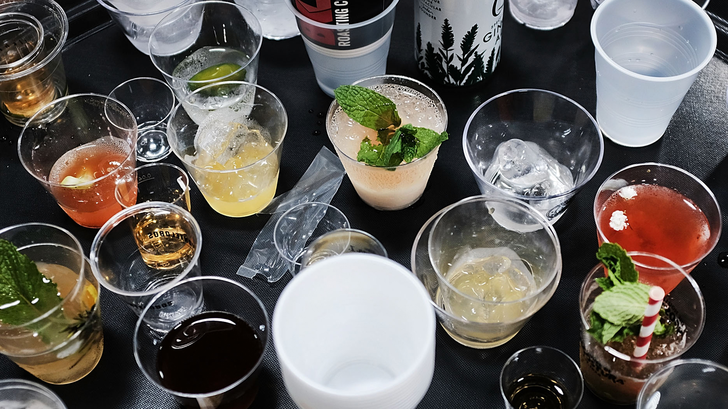 An assortment of half-empty drinks in various plastic cups, some with ice and garnishes, is spread across a dark surface—a telltale sign of the previous night's hangover remedy attempts.