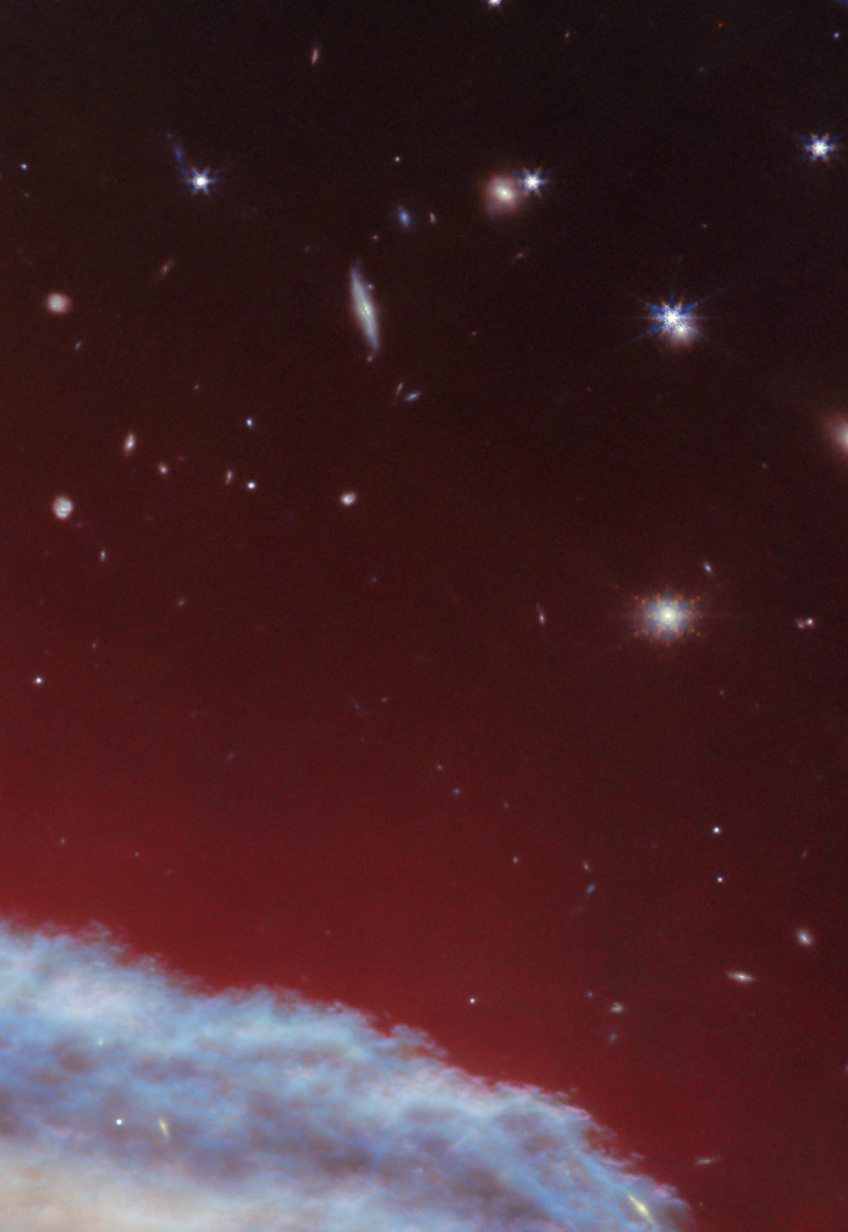 A vibrant image captured by the JWST of space featuring a blue nebulous cloud at the bottom with a scattering of stars and distant galaxies against a deep red backdrop.