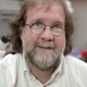 A middle-aged man with a beard and glasses, wearing a light yellow shirt, smiling in an indoor setting.