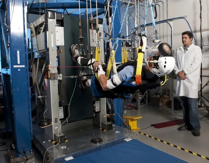 A person is suspended in a complex harness system and wearing a helmet, while a scientist in a lab coat observes. The setup appears to be part of an experimental or training apparatus.