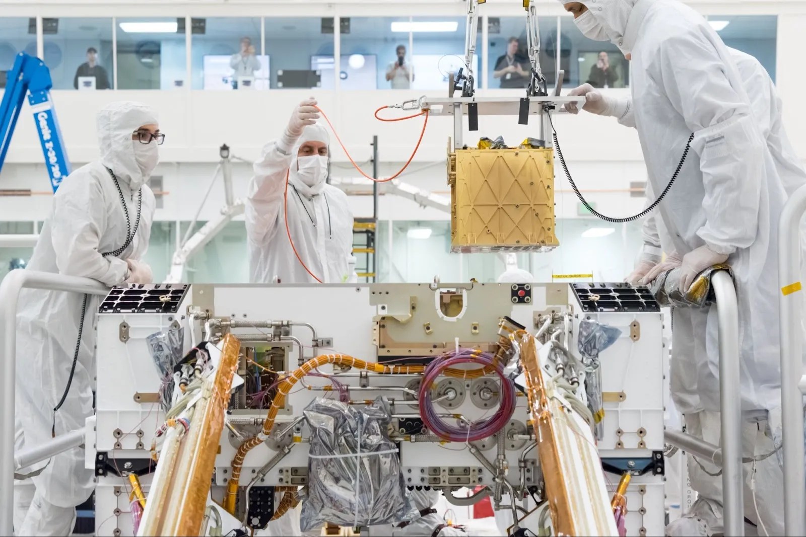 Scientists in white protective suits work on assembling a complex machine in a cleanroom environment, with various technical instruments and wires visible.