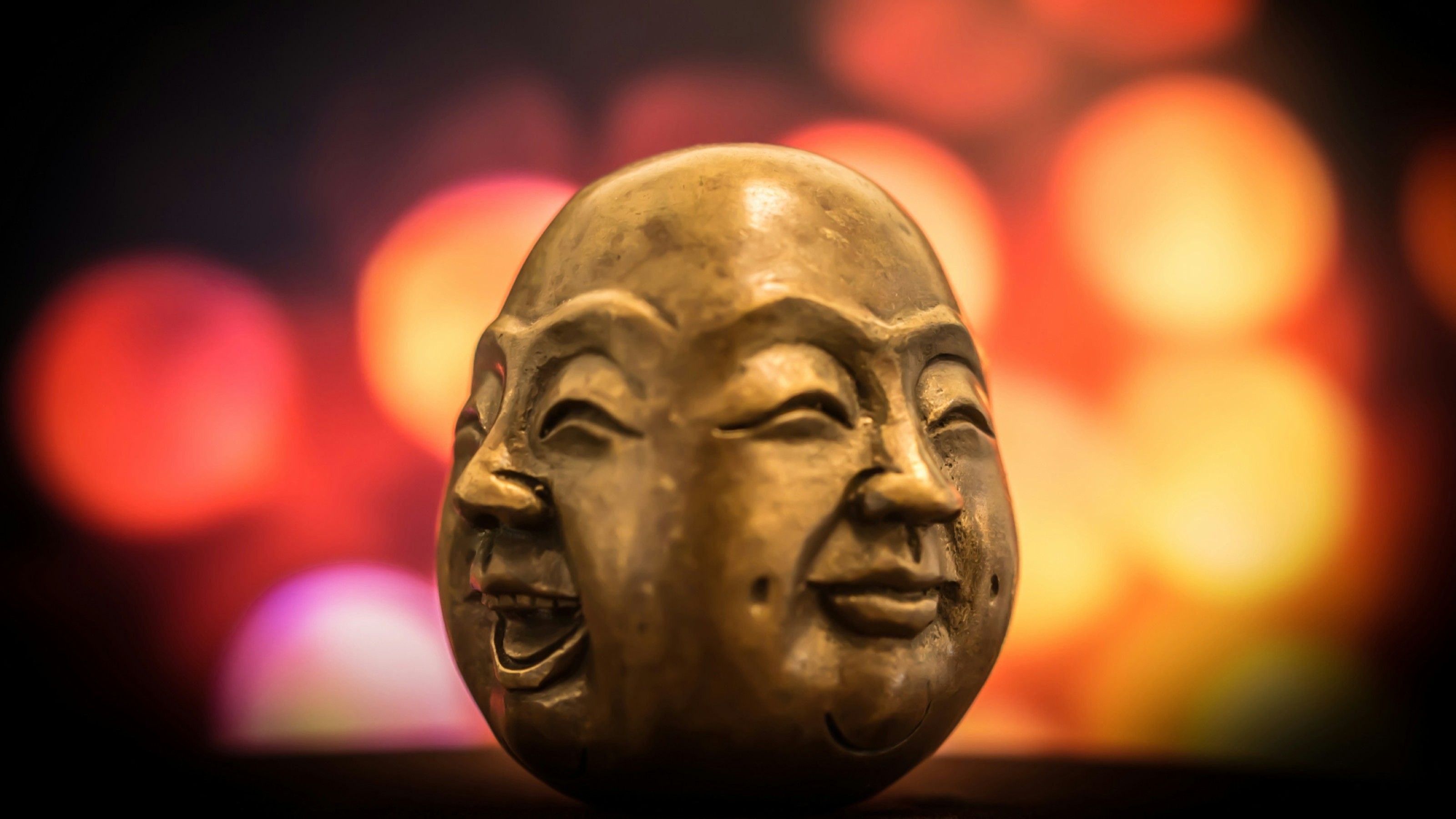 A small statue depicting a face with dual expressions, smiling and neutral, against a blurred background of colorful bokeh lights.
