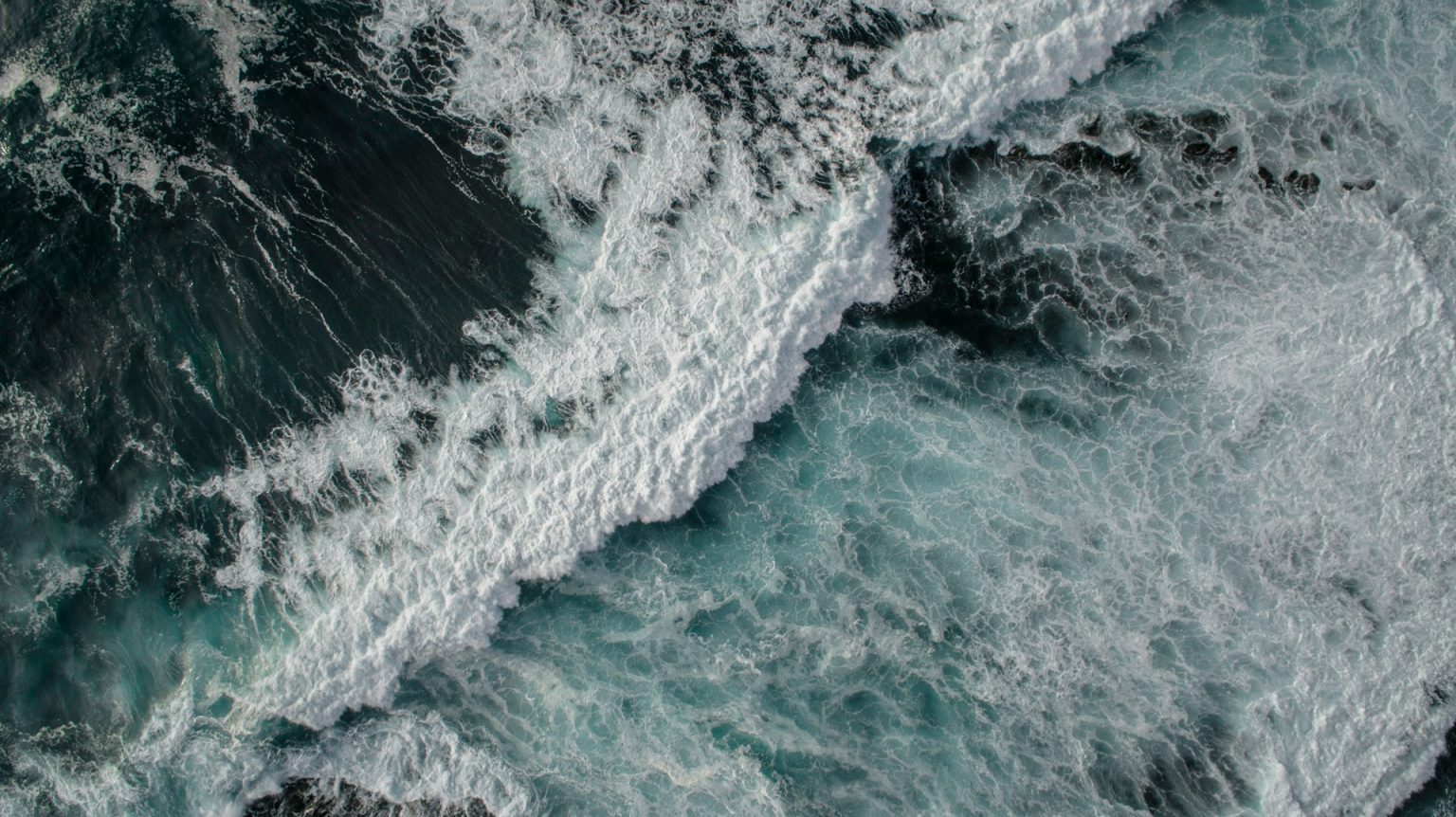 Aerial view of ocean waves crashing and foaming, creating a turbulent pattern in the dark and light blue water.