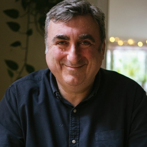 A middle-aged man with gray hair and a slight smile, wearing a dark button-up shirt, is sitting indoors with a plant and some string lights in the background.