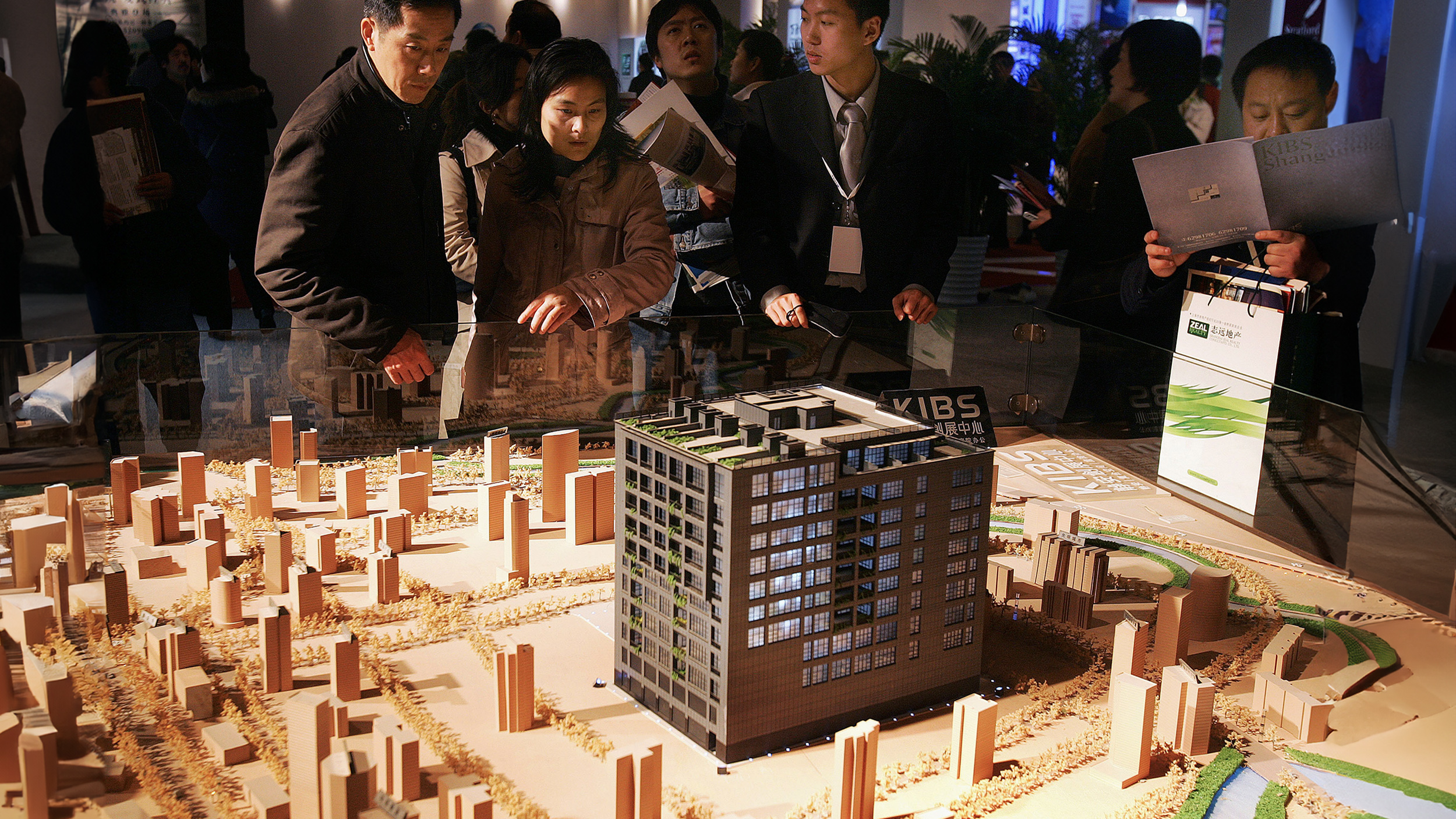 People examining a detailed architectural model of a building and its surrounding area at an exhibition, reflecting the impact of the Chinese economy on urban development.