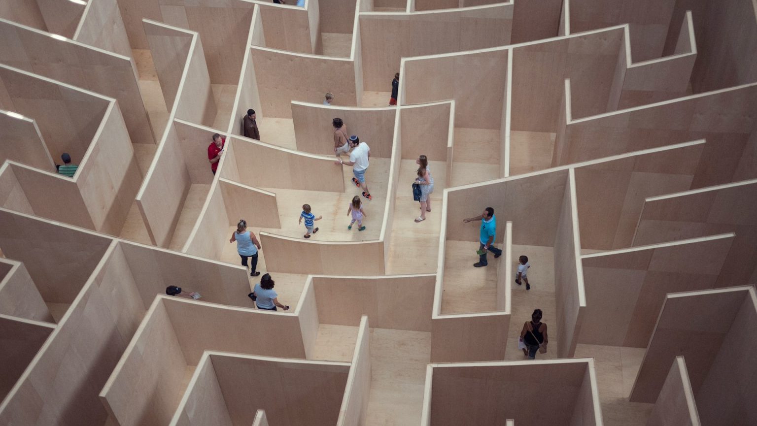 Visitors exploring why people get lost in a large, complex wooden maze installation in an indoor gallery setting.