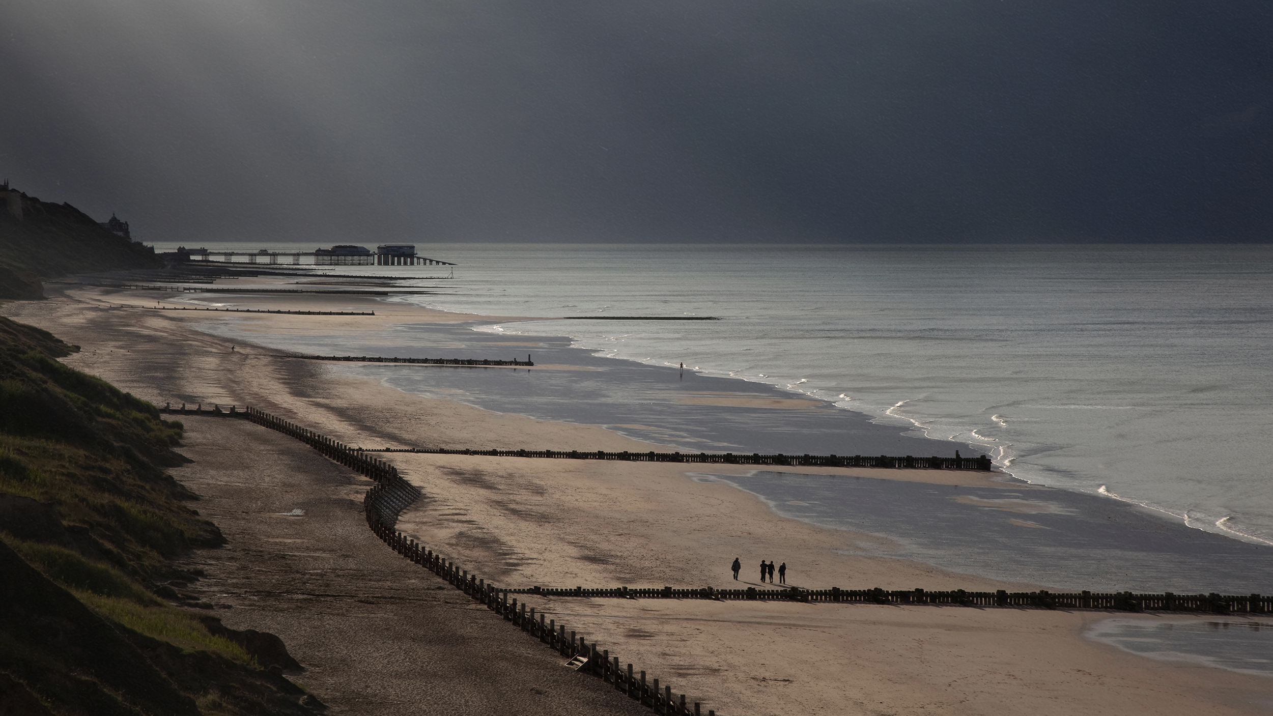A serene beach scene at dusk with a distant pier, a few people walking along the shoreline, and gentle waves hitting the sand. The sky is cloudy with rays of light breaking through, hinting at the subtle potential for tidal power beneath the surface.