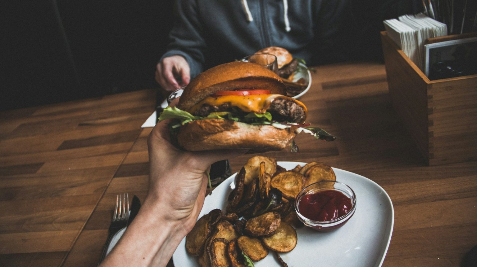 A person holding a burger with fries and ketchup on a wooden table.