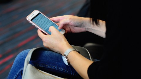 Person seated, using a smartphone to browse social media, with a focus on their hands and the device over a handbag, inside a room with striped carpeting.