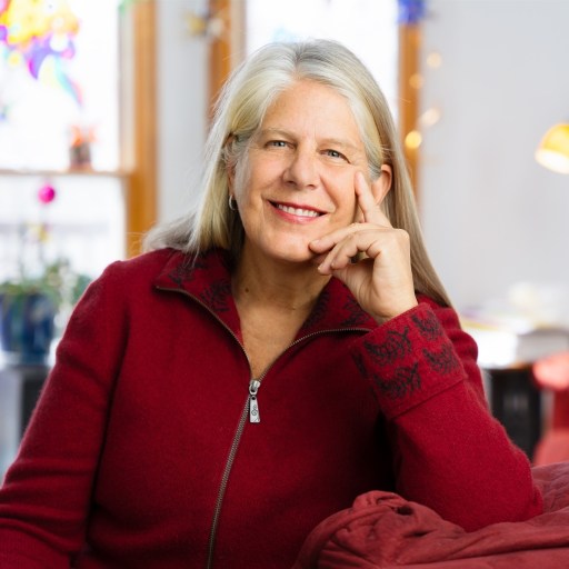 A woman with long gray hair wearing a red zip-up sweater sits in a brightly lit room, smiling with her head resting on her hand.