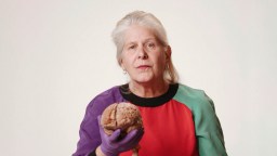 A woman with grey hair, wearing a colorful sweater and purple gloves, holds a human brain.