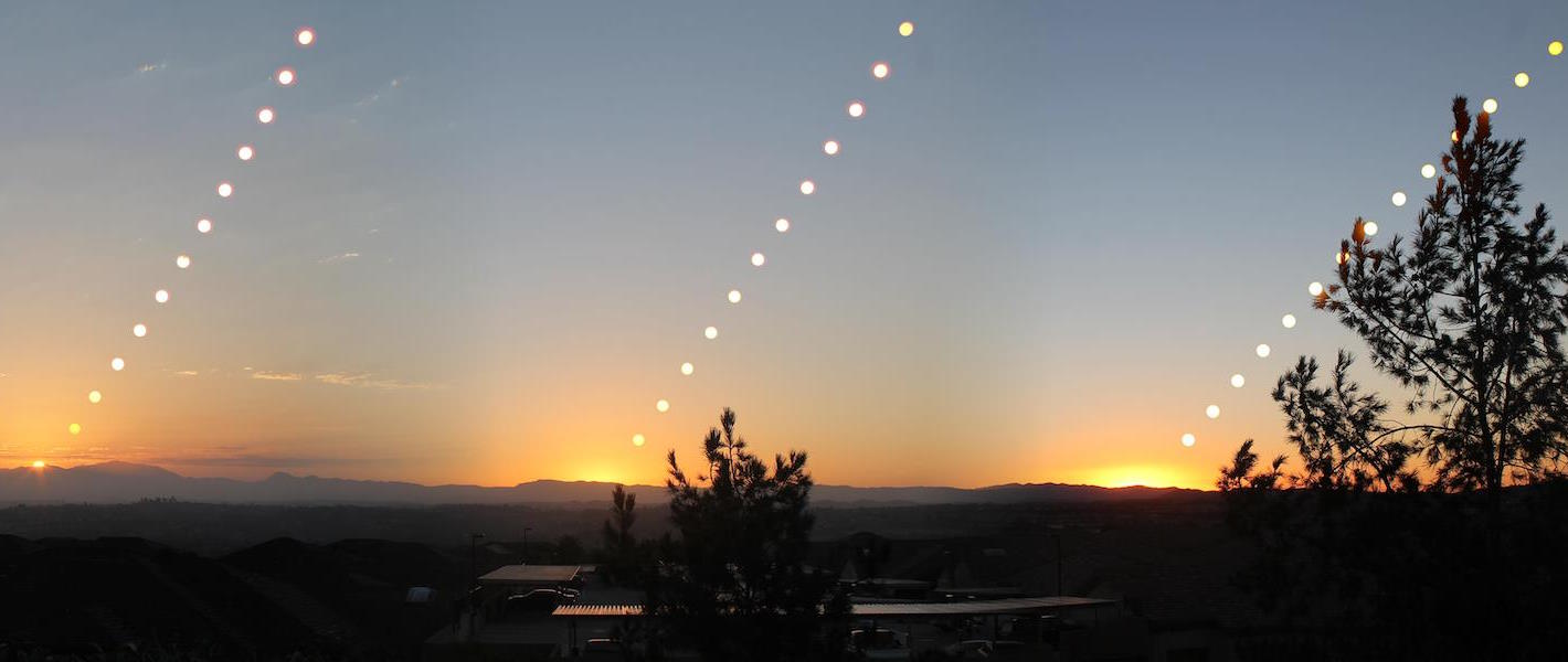 A series of sun positions during sunset over a landscape, with trees in the foreground and mountains in the background, creating a pattern of glowing points in the sky.