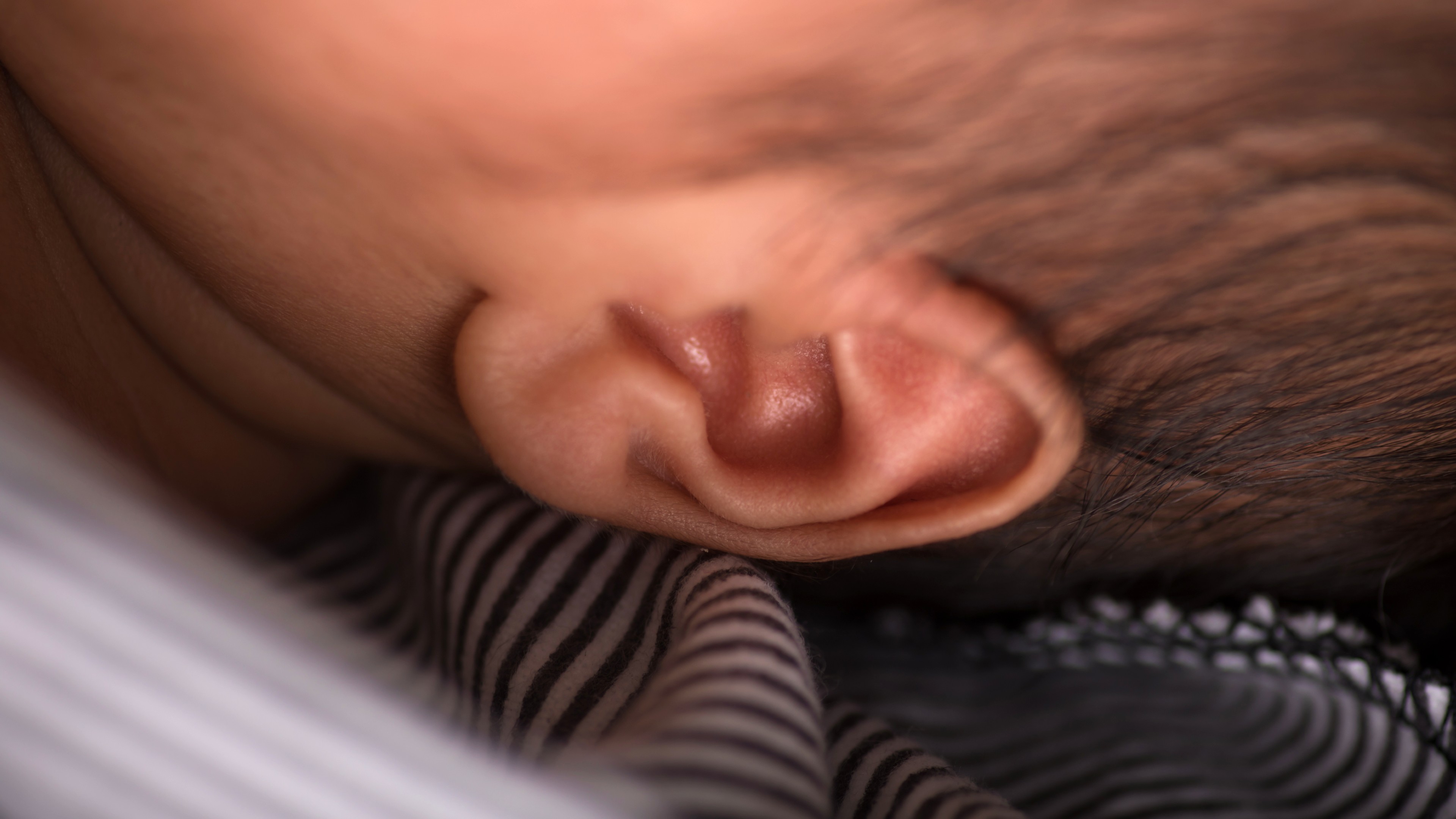 Close-up image of a baby's ear as the baby rests its head against a striped fabric, capturing the tender moments when they first start to absorb the language around them.