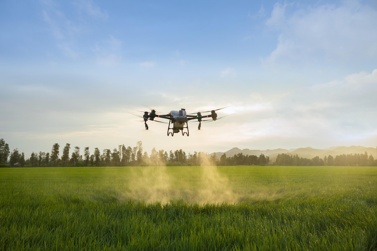 A drone flying over a green field sprays pesticide, with trees and mountains in the background under a blue sky.