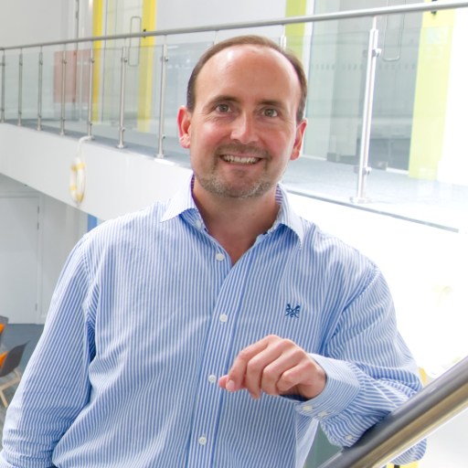 A man in a blue striped shirt and black pants stands leaning on a railing indoors, smiling at the camera.